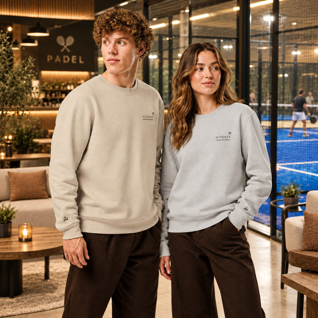Padel Tennis Pullover für Damen und Herren, Sweater für Padel Tennis Spieler, Mann und Frau stehen im Padel Outfit in einer Padel Lounge. Im Hinterrund spielen Menschen Padel Tennis auf einem Indoorplatz.